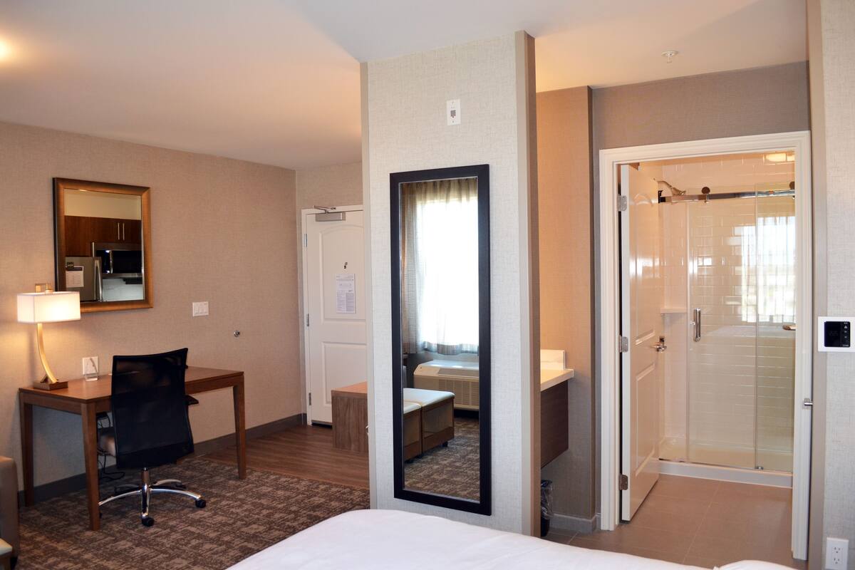A hotel room with a bed, desk, chair, mirror, and a shower area.