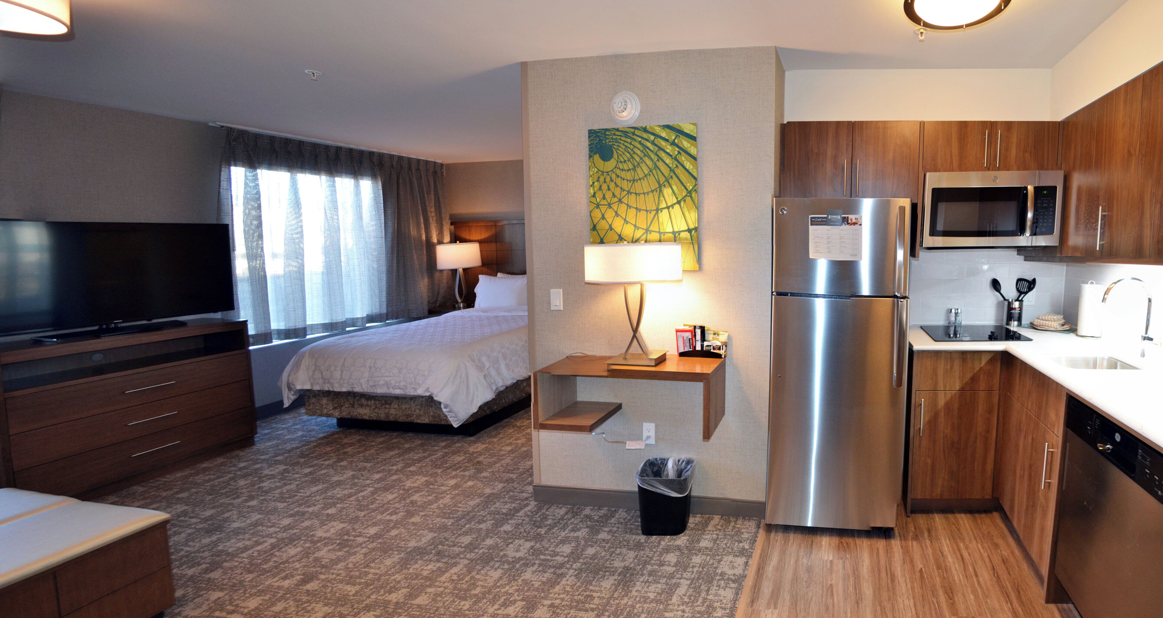 A hotel room with a bed, a TV, a desk with a lamp, a refrigerator, a microwave, and a sink.