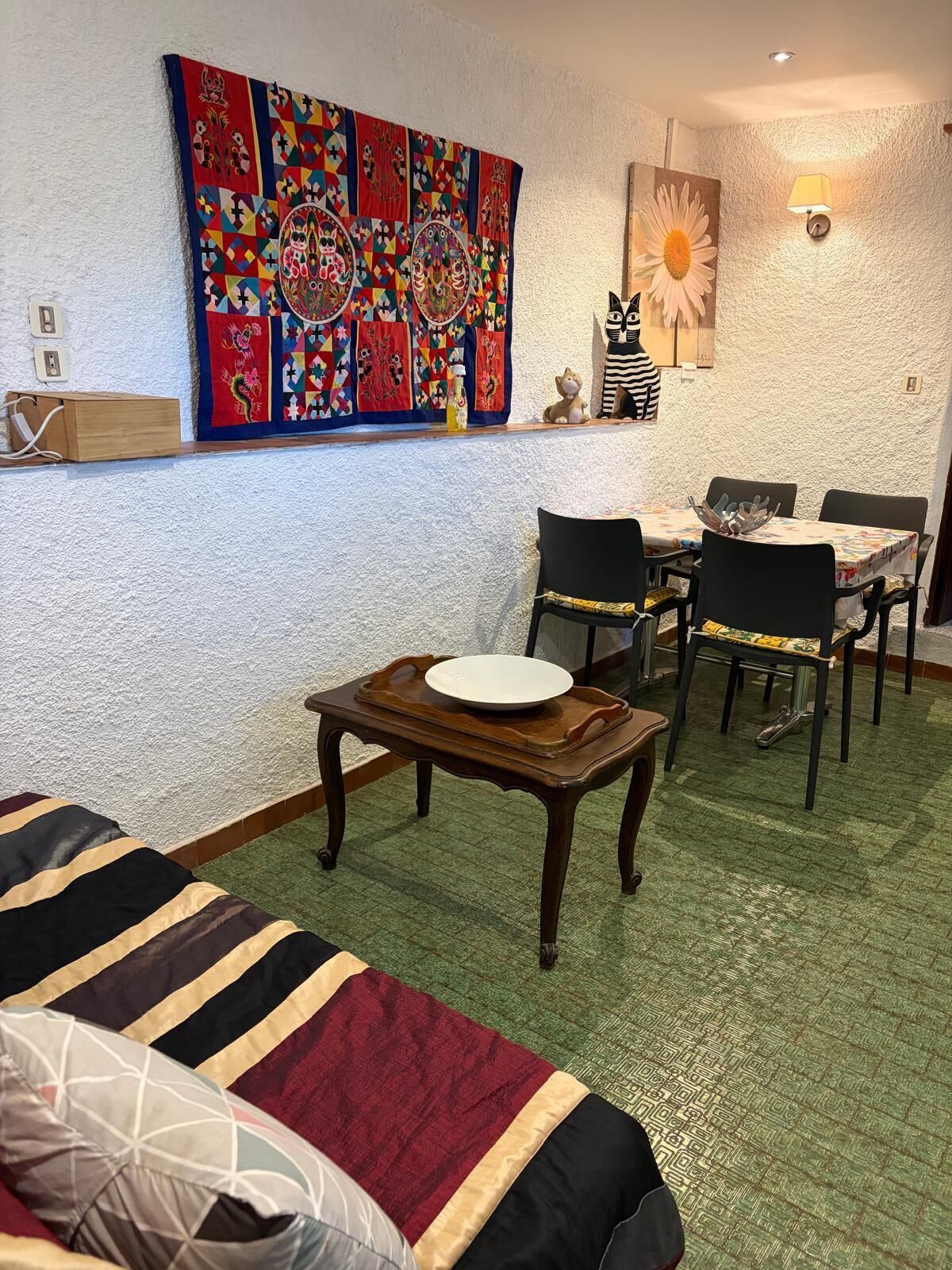 A room with a green carpet, a sofa, a dining table with chairs, a wall with a colorful quilt, and a wall-mounted lamp.