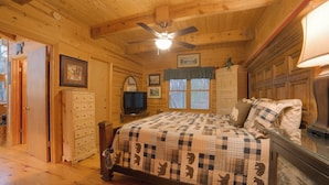 A wooden cabin bedroom with a bed, dresser, television, and a ceiling fan.