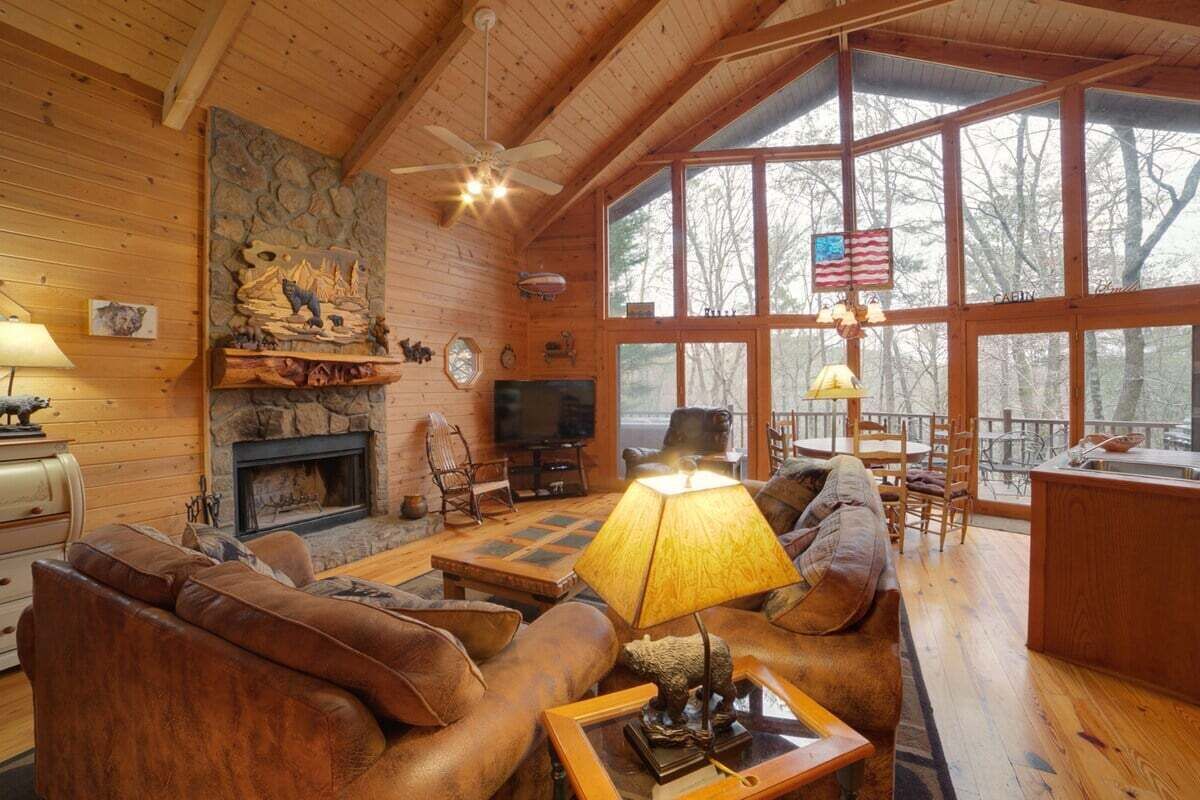 A rustic cabin interior with a fireplace, leather sofa, wooden floors, and large windows offering a view of the surrounding forest.