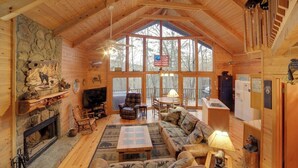 A cozy log cabin interior with a stone fireplace, a large sofa, a television, and a dining area.