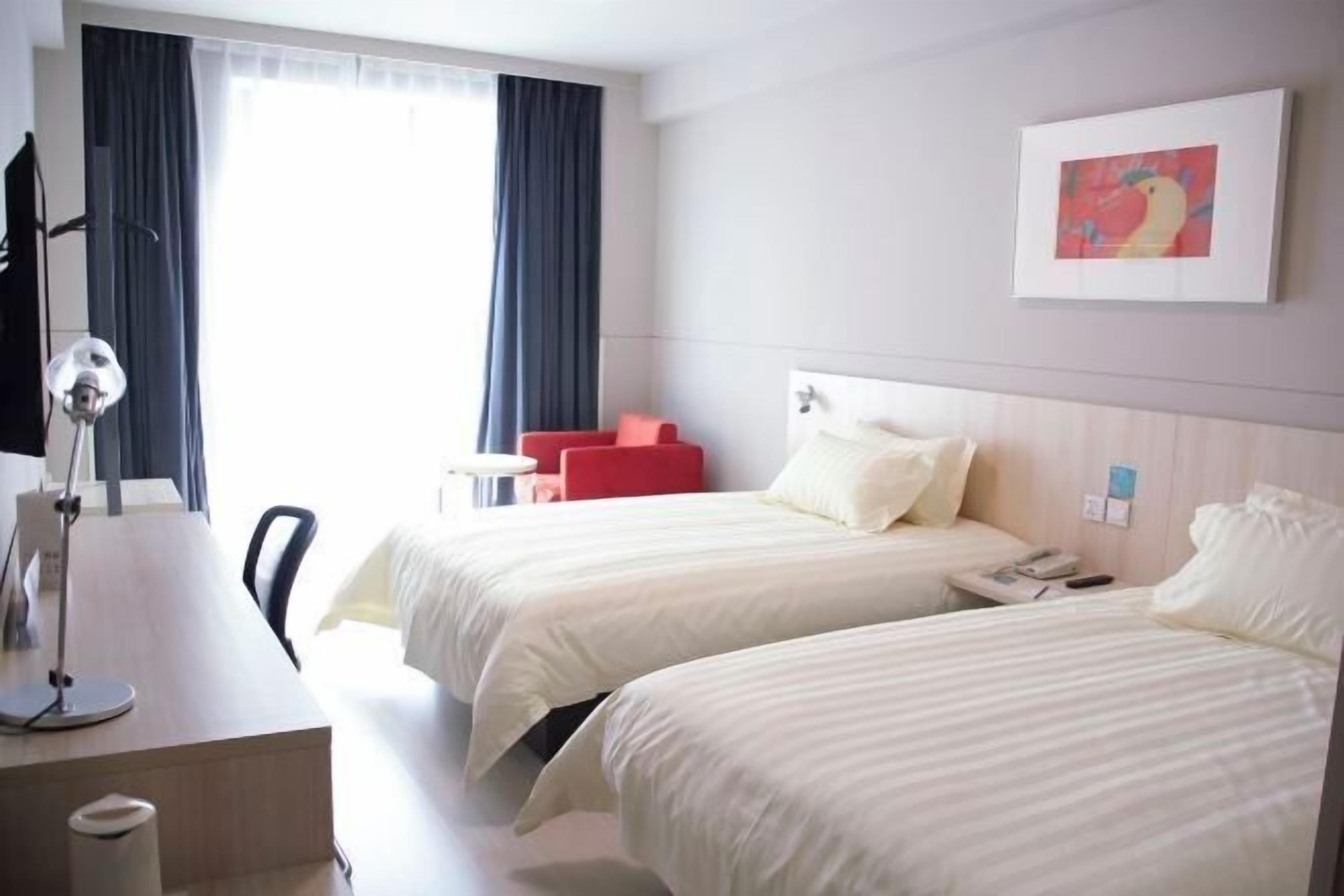Habitación de hotel con dos camas, un escritorio, una silla, una lámpara, una butaca roja, un cuadro y una ventana con cortinas.