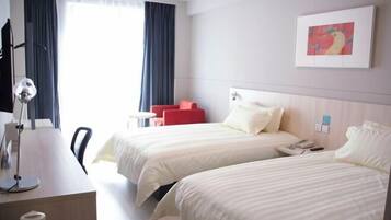 Habitación de hotel con dos camas, un escritorio, una silla, una lámpara, una butaca roja, un cuadro y una ventana con cortinas.