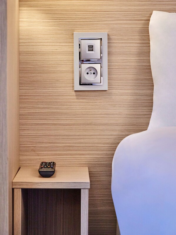 Une chambre d’hôtel moderne équipée d’une prise électrique intégrée et d’une télécommande posée sur une table de chevet.