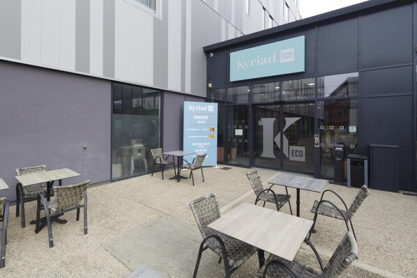 Un espace extérieur aménagé avec des tables et des chaises, un comptoir d’accueil et une enseigne indiquant « Kyriad ECO ».