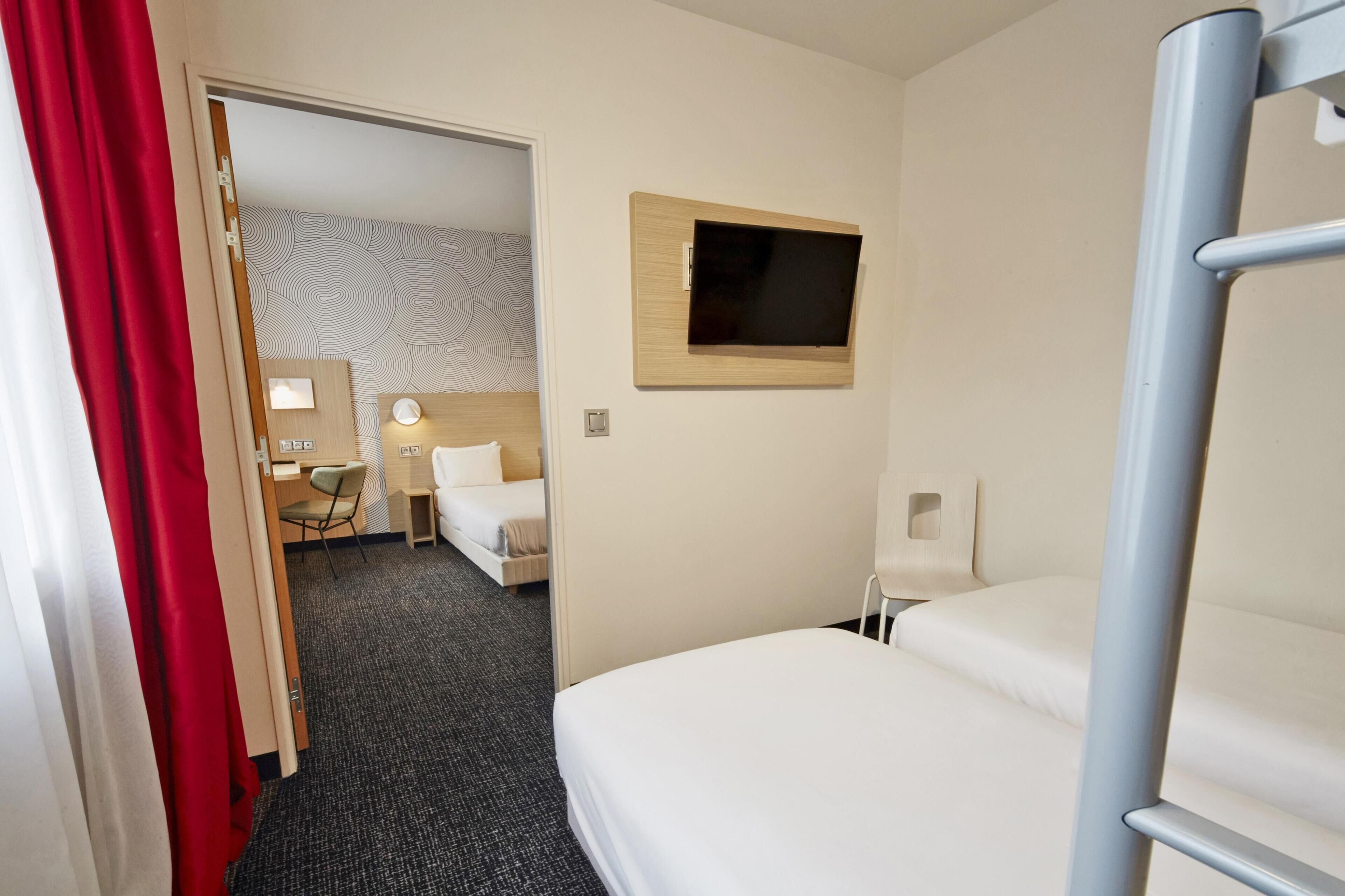 A hotel room with a bed, a desk, a chair, a TV mounted on the wall, and a door leading to another room.
