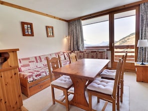 Uma mesa de jantar de madeira com cadeiras estofadas, um banco com tecido estampado e vista para uma paisagem montanhosa através de grandes janelas.
