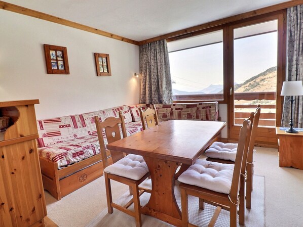 A wooden dining table with cushioned chairs, a bench with patterned upholstery, and a view of a mountainous landscape through large windows. - Charming 2-Room Apartment for 4 - Comfort and Wi-Fi (Méribel)