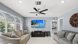 A modern living room with a large flat-screen TV, two sofas, a ceiling fan, and a large wall clock. - Stunning Home in Beautiful "Floresta Pines" Port St Lucie! (Port St. Lucie)