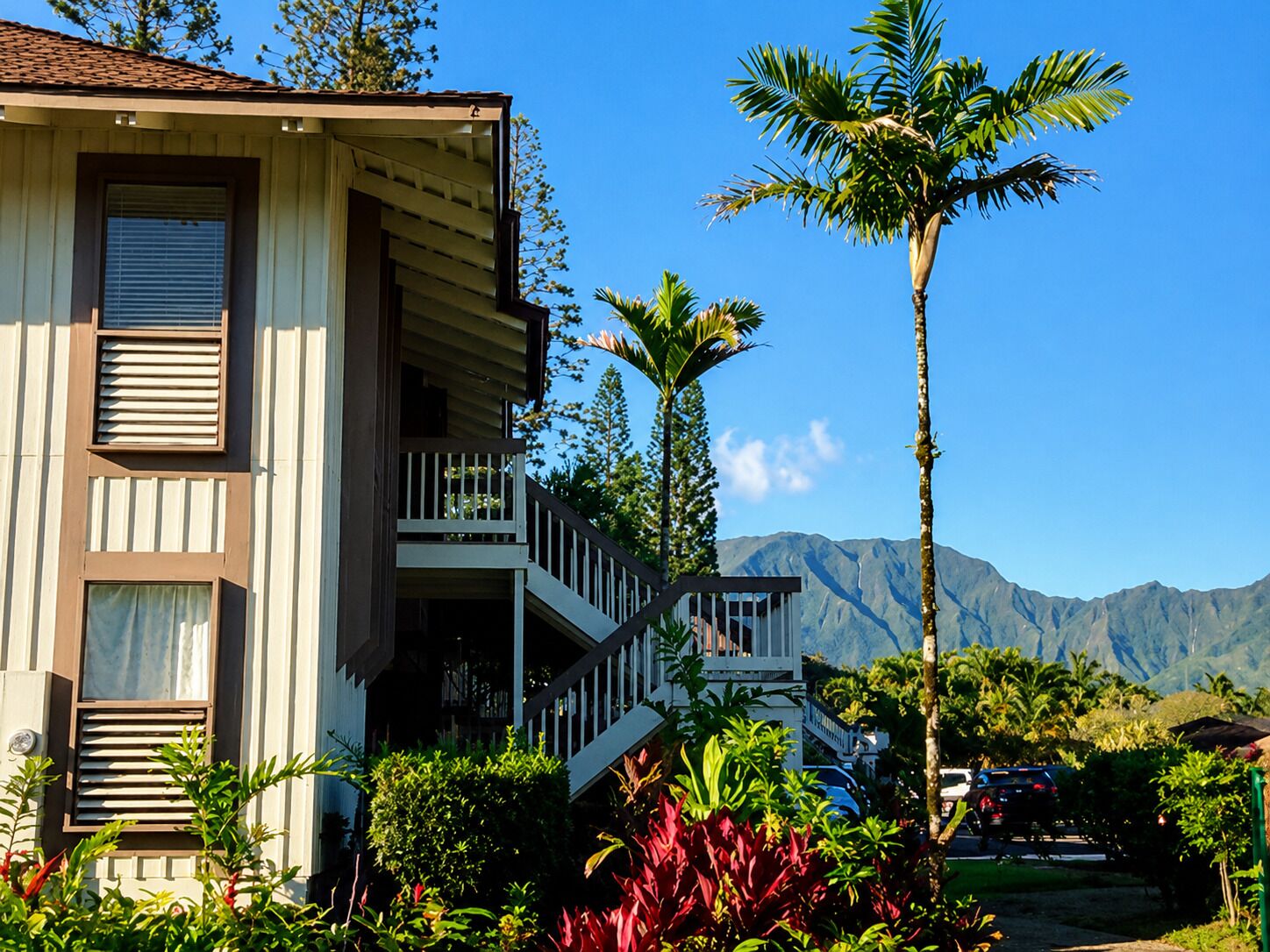 A two-story house with a balcony, surrounded by tropical vegetation and mountains in the background.