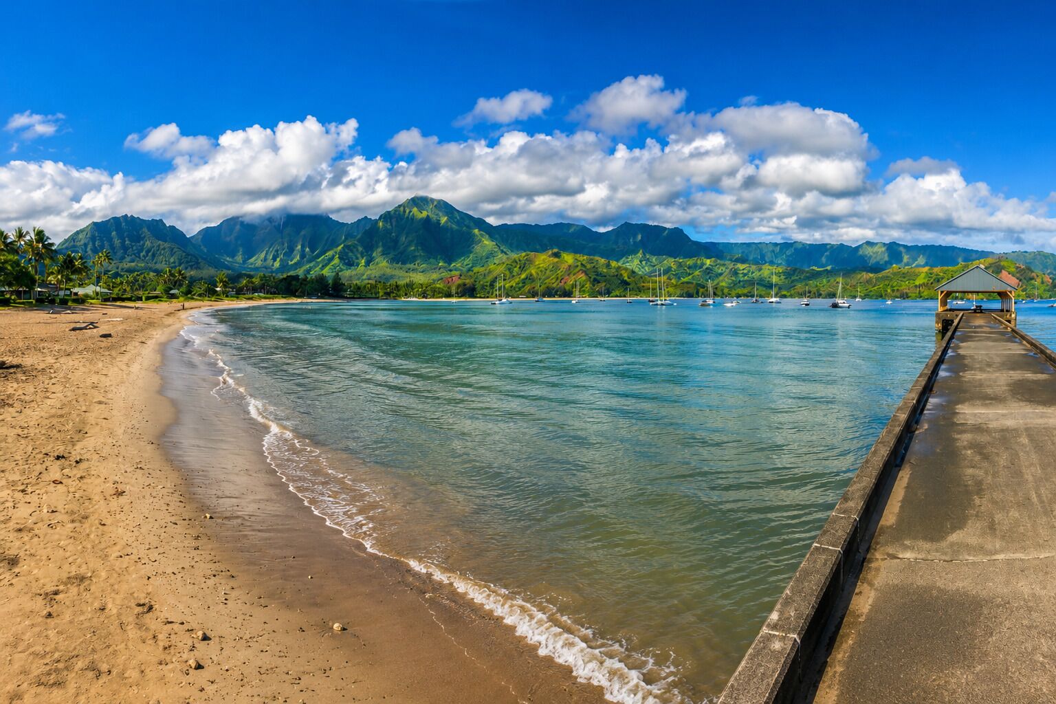 A beach with a concrete pier extending into calm waters, surrounded by lush green mountains and clear skies.