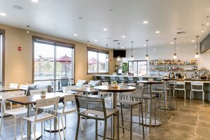A spacious cafe with multiple seating areas, a bar, and large windows offering views of the outside.
