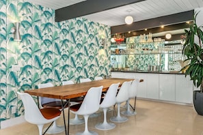 Un comedor con una mesa de madera y sillas blancas, una cocina con armarios de vidrio y una pared con estampado de hojas tropicales.