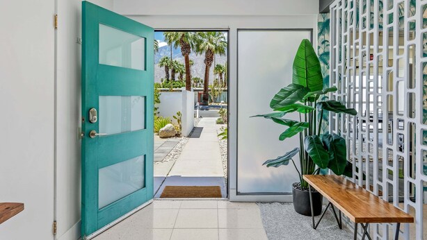 Un pasillo con una puerta verde azulada, un banco de madera, una planta en maceta y un separador decorativo blanco.