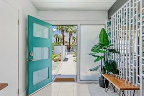 Un pasillo con una puerta verde azulada, un banco de madera, una planta en maceta y un separador decorativo blanco.