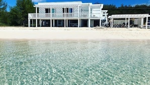 En fastighet som ligger vid stranden, med en vit byggnad, klart och grunt vatten samt en sandstrand.