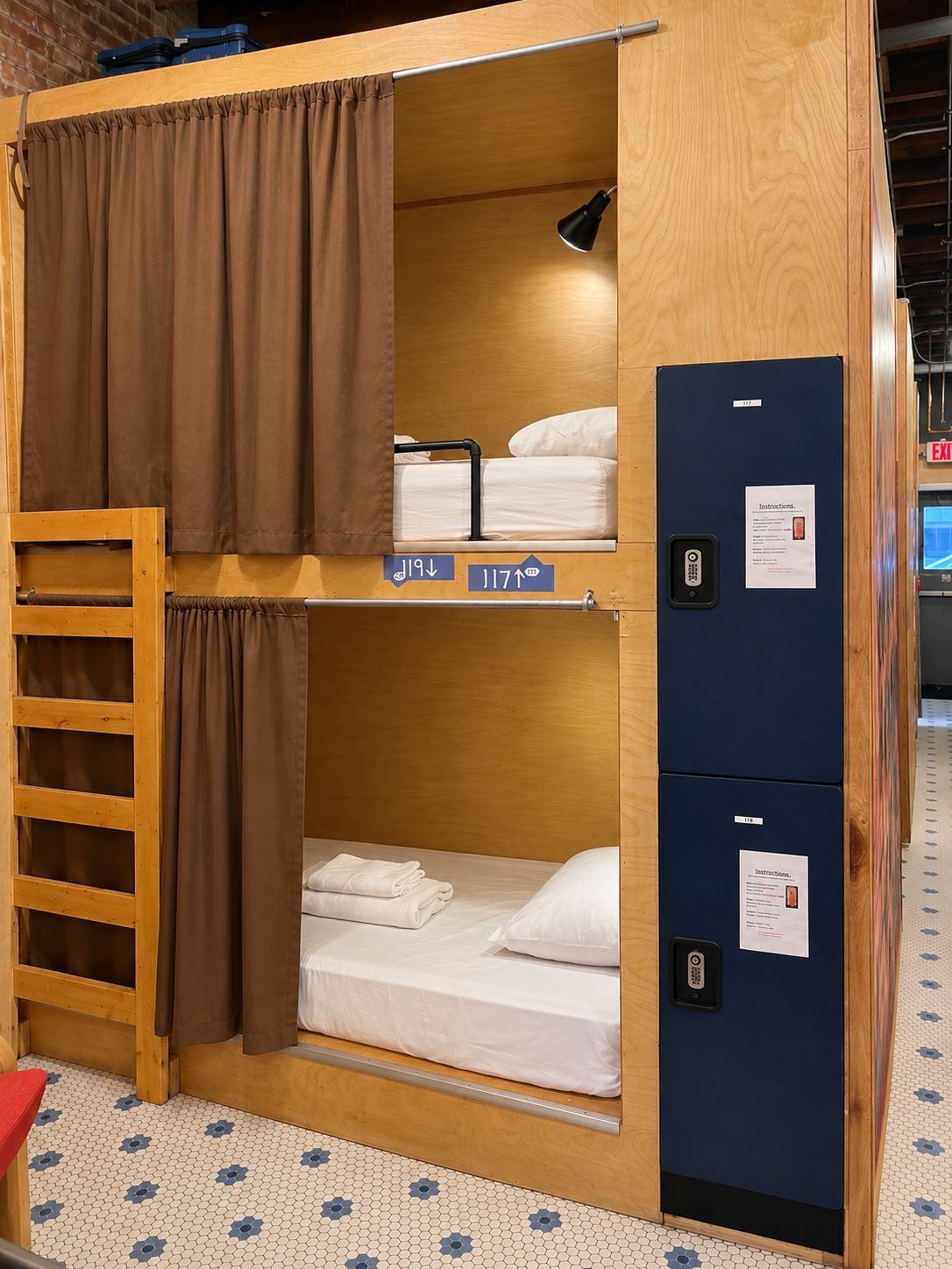 A bunk bed room with a ladder, a desk, and a computer. There are signs indicating room numbers and a door with an exit sign.