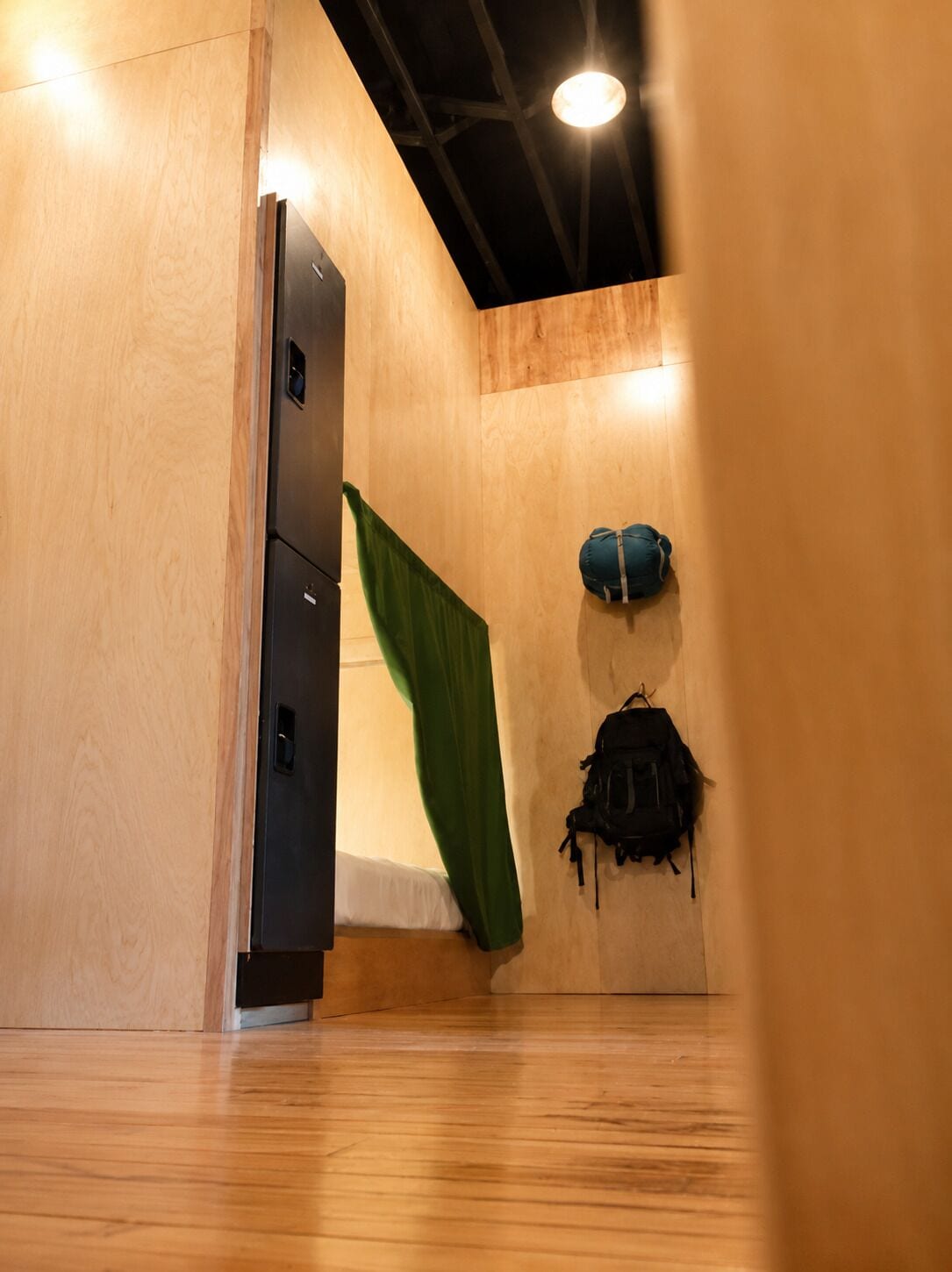 A room with a wooden floor, a bed with a green curtain, a black backpack, and a black cabinet.