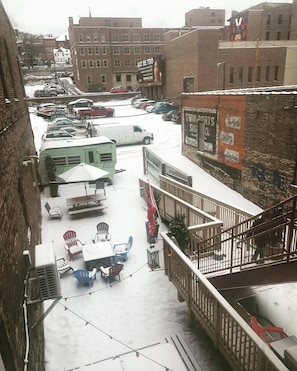 A snowy urban outdoor area with parked cars, a green RV, outdoor seating, and a staircase leading to a building with a mural.