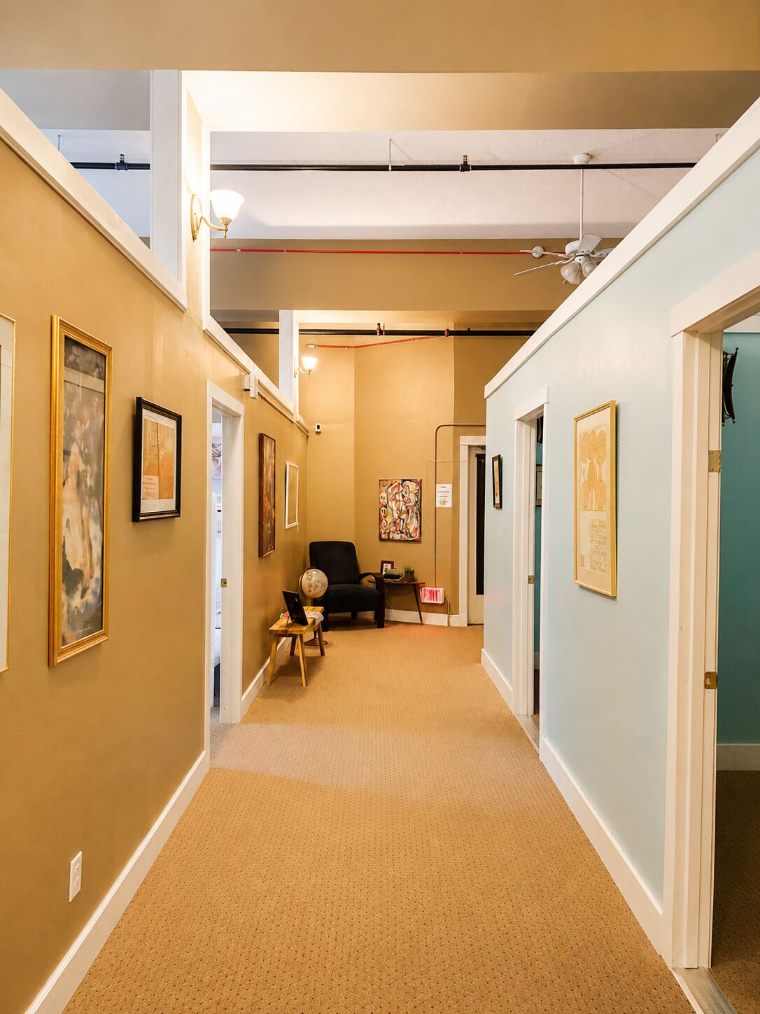 A hallway with beige walls, framed artwork, a black chair, and a desk with a laptop.