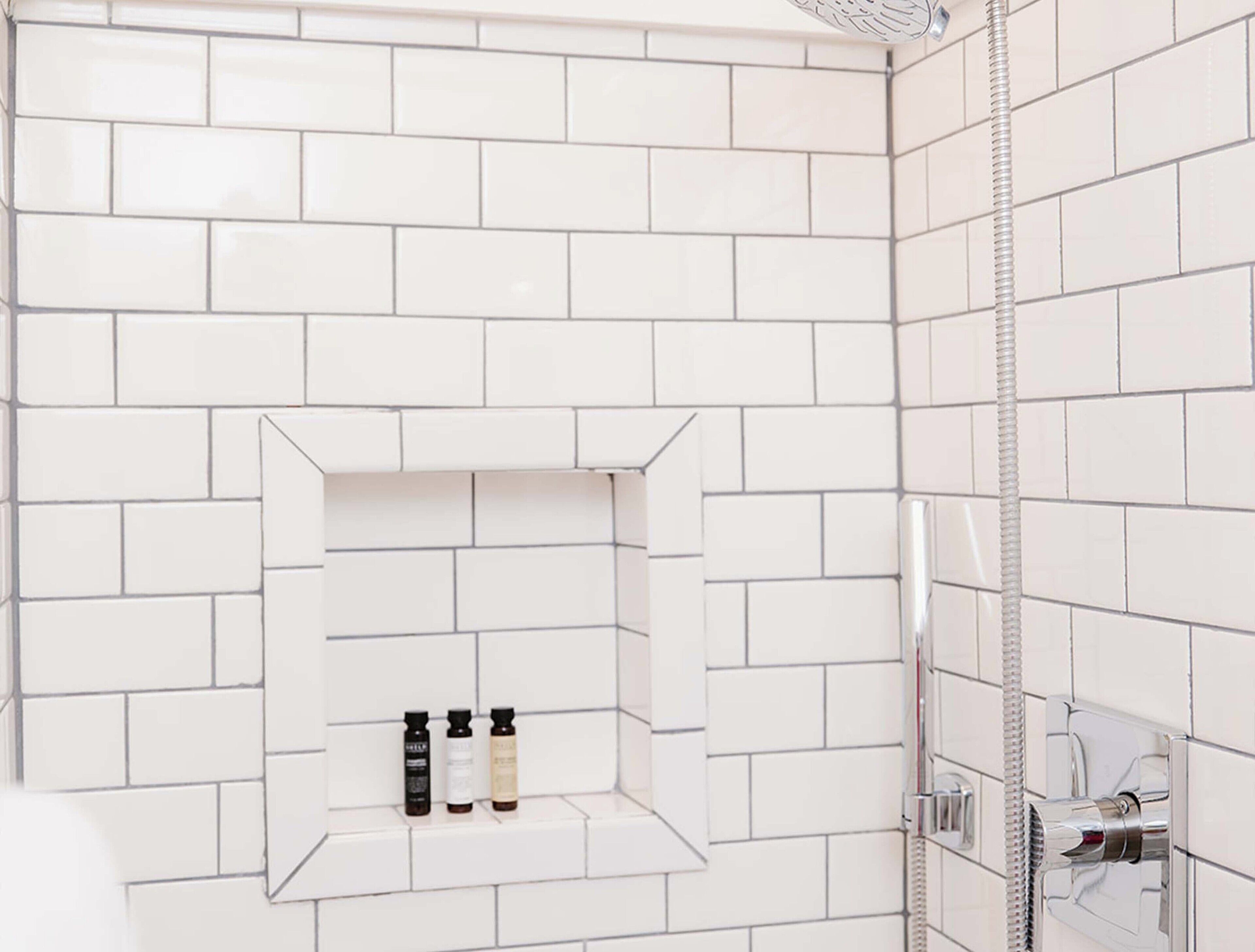 Zona de ducha con azulejos blancos, un estante empotrado con tres botellas y un cabezal de ducha.