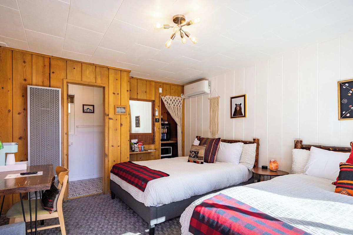 A cozy room with two beds, a desk, and a chair. There is a window with curtains, a ceiling fan, and a wall-mounted air conditioner.