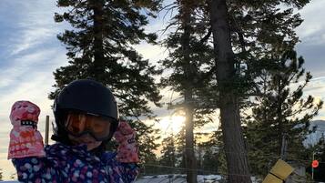 A child in a blue and pink snowsuit standing on a sled in a snowy area with trees and a sunset in the background.
