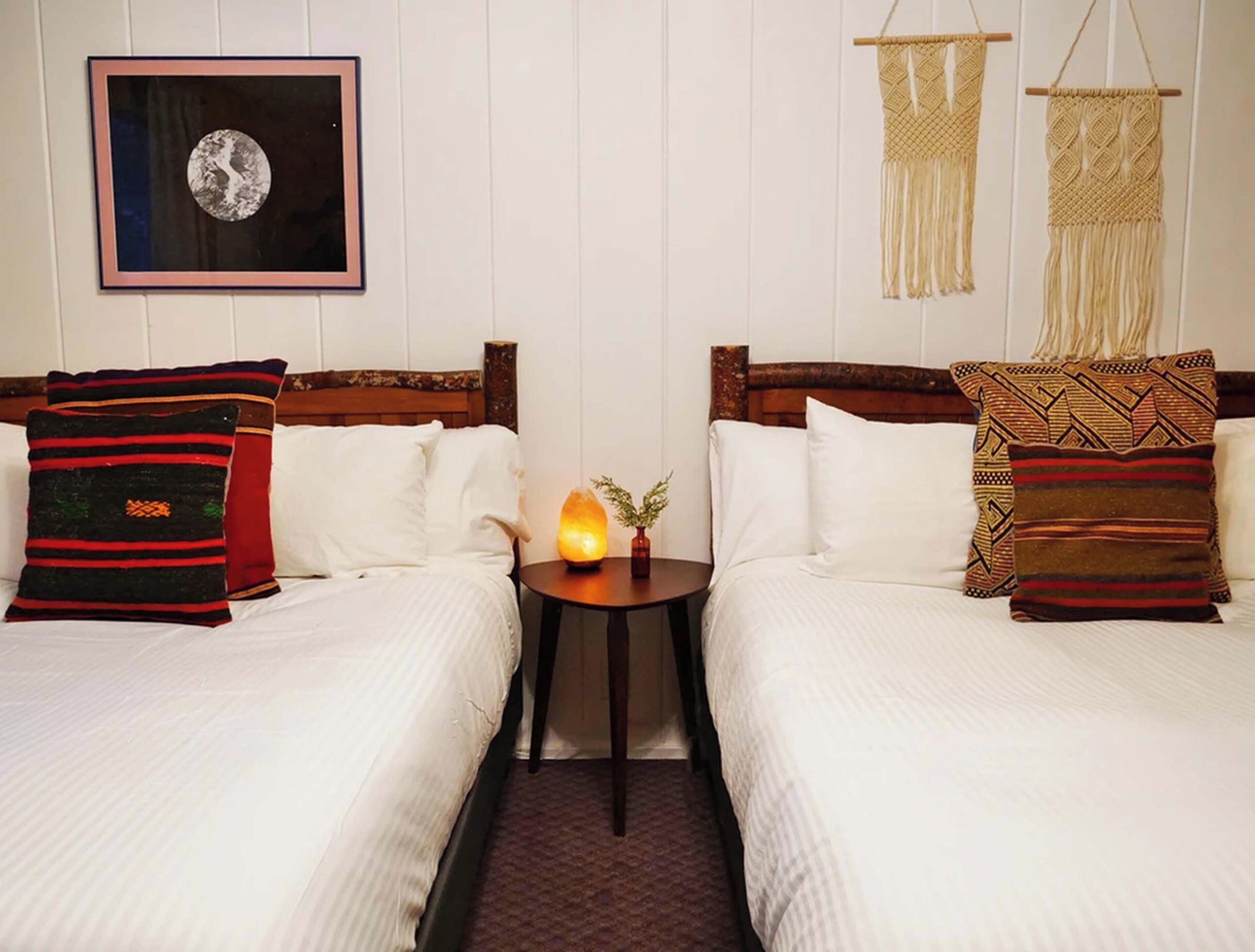A hotel room with two beds, a framed picture on the wall, a small table with a heater, and macramé wall hangings.