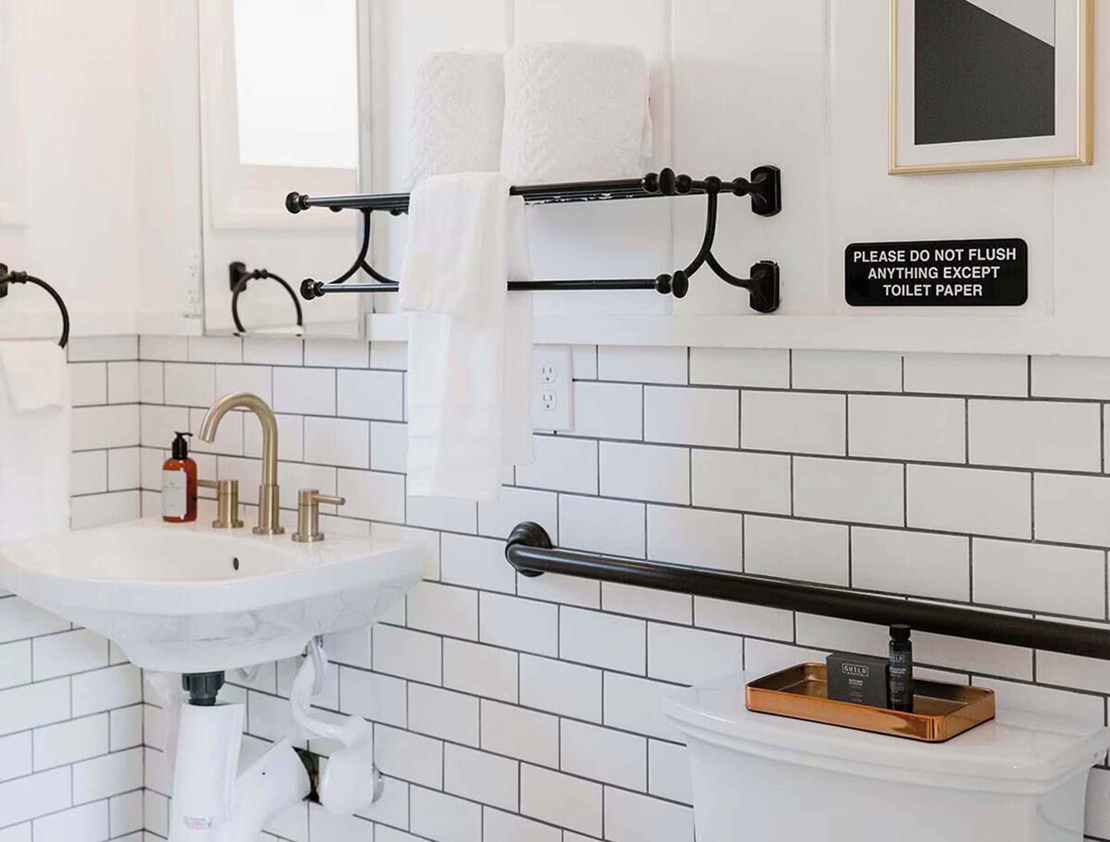 A bathroom with white tiled walls, a white sink with gold faucets, and a toilet with a black grab bar. There is a sign on the wall that reads "PLEASE DO NOT FLUSH ANYTHING EXCEPT TOILET PAPER."