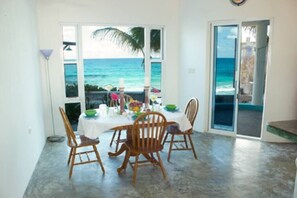 Comedor con vista al océano, una mesa blanca, sillas de madera y puerta de vidrio que da al exterior.