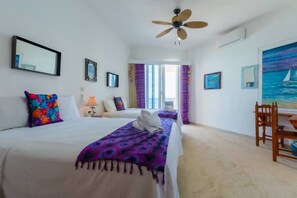 Una habitación de hotel con dos camas, un ventilador de techo, una zona de comedor con mesa y sillas, y un cuadro de un velero en la pared.