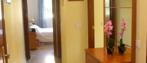 Un pasillo de hotel con una cama al fondo, un aparador de madera con espejo y una pequeña planta en maceta.