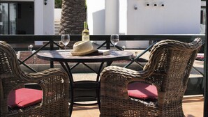 Una mesa con dos sillas de mimbre, una botella de vino y una copa en una terraza con vistas a un edificio blanco y una palmera.