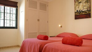 Un dormitorio con dos camas, cada una con una manta roja y una toalla enrollada. Hay una ventana con vistas a árboles, un cuadro en la pared y un interruptor de luz.