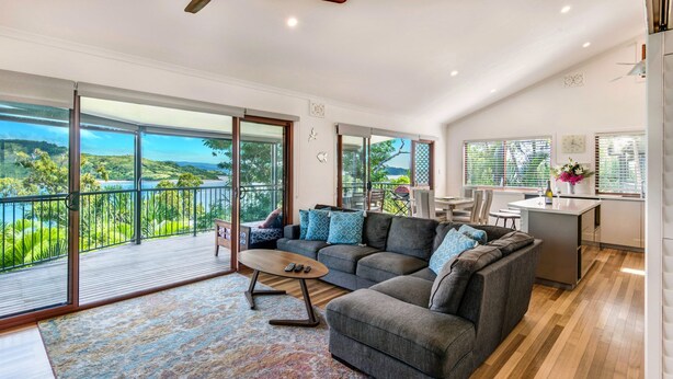 A spacious living area with a large gray sofa, a coffee table, and a view of a lush landscape through expansive glass doors. - Heliconia 10 Hamilton Island - oceanviews, buggy and transfers (Hamilton Island)