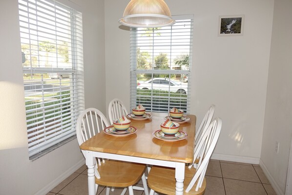 A dining area with a wooden table and white chairs, a hanging light fixture, and a window with blinds. - BrightwaterVilla. Lake view, west facing extra large pool deck, lanai, hot tub (Kissimmee)