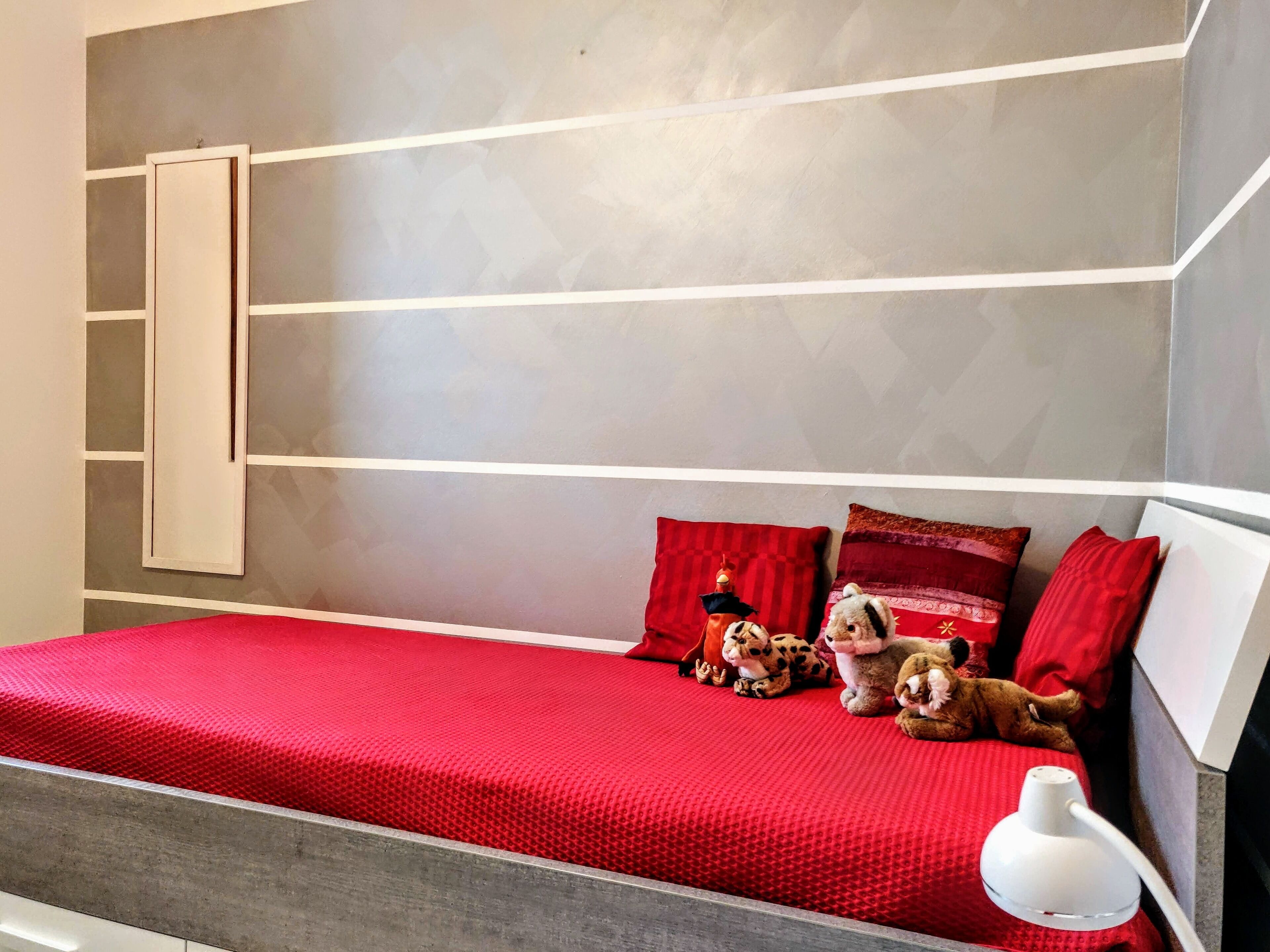 A modern bedroom with a red bedspread, a white bedside lamp, and plush toys on the bed.