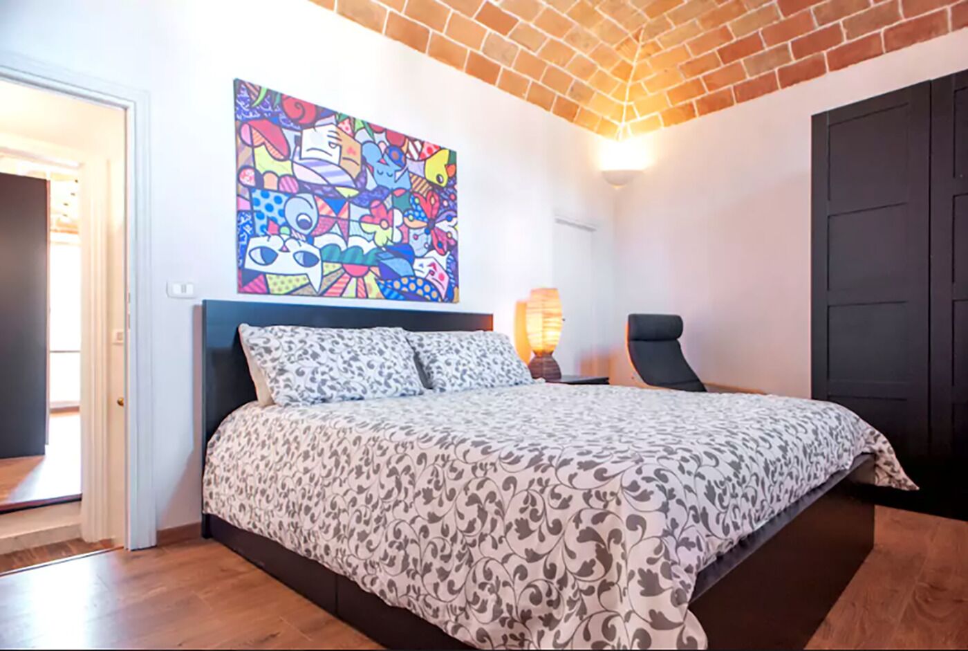 Une chambre avec une couvre-lit à motifs, un tableau abstrait coloré au-dessus du lit, une chaise, une table de chevet avec une lampe et une porte.
