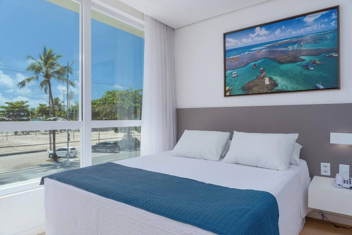 Habitación de hotel con una cama grande, un cuadro de una escena costera y vistas a la calle y palmeras por la ventana.