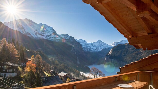 A wooden deck with a table and chairs overlooking a mountainous landscape with a lake and snow-capped peaks. - 5 Star Penthouse Apartment in Chalet Aberot !! (Wengen)