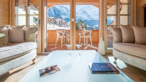 A cozy room with a sofa, a coffee table with a book and glasses, and a balcony with a mountain view. - 5 Star Penthouse Apartment in Chalet Aberot !! (Wengen)