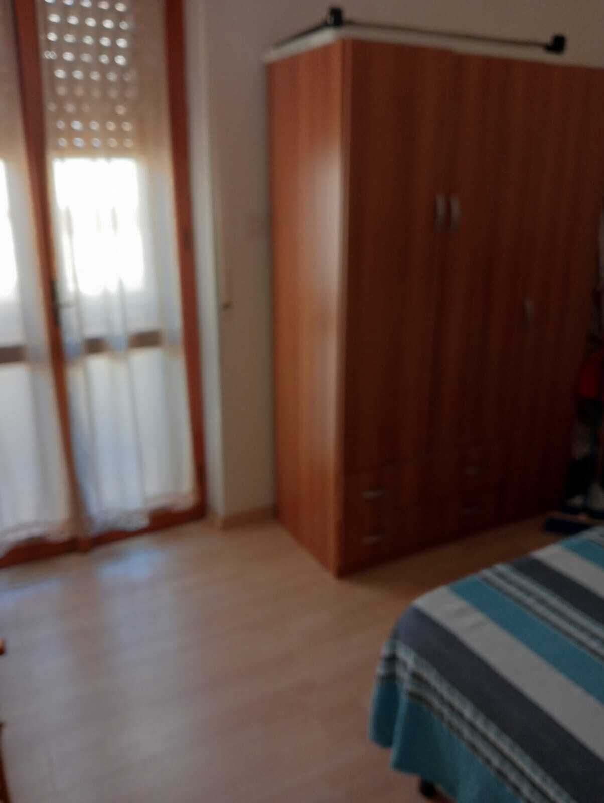 Ein Schlafzimmer mit einem Holzschrank, einem Bett mit gestreifter Bettdecke und einem Fenster mit transparenten Vorhängen.