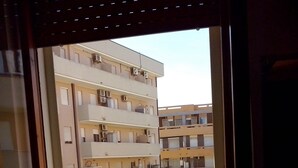 A balcony with a potted plant, a view of a multi-story building, and a car parked on the street.