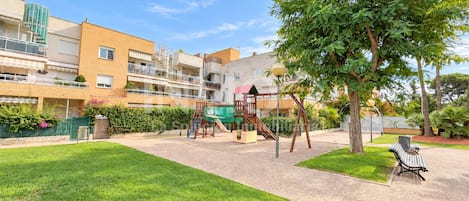 A residential building with a playground, green lawn, and a bench.