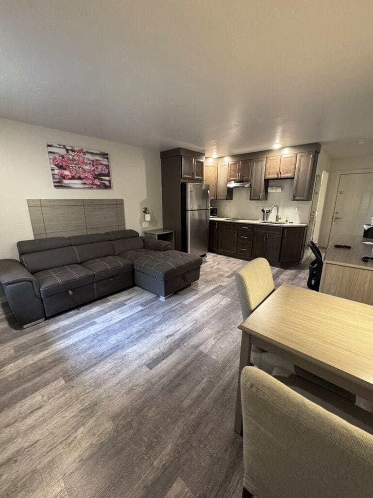 A modern living room with a sectional sofa, a dining table, and a kitchen area with dark cabinets and a refrigerator.