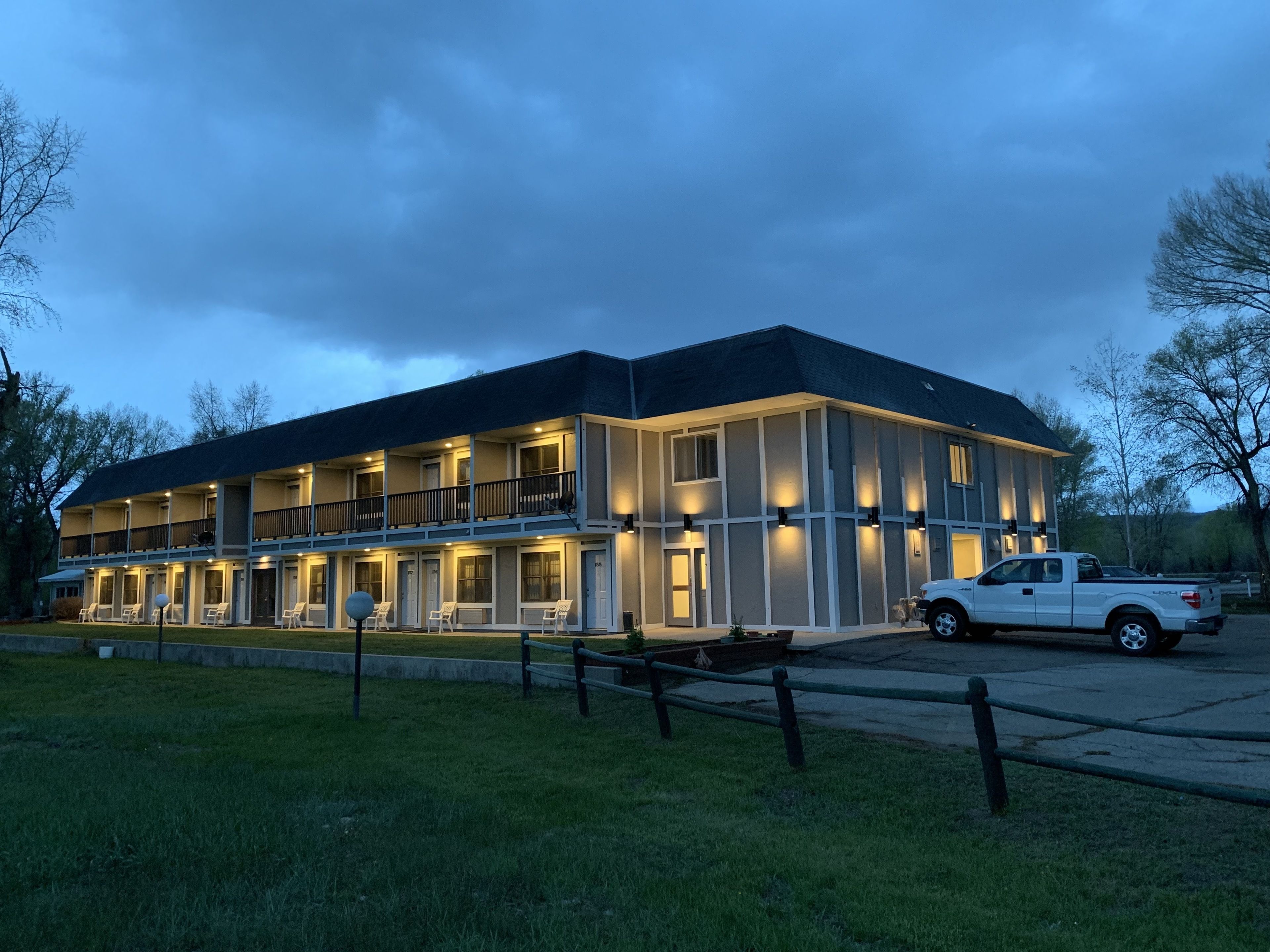 Un motel de varios pisos con una hilera de habitaciones en la planta baja, un área de patio techada y una camioneta pickup blanca estacionada al frente.