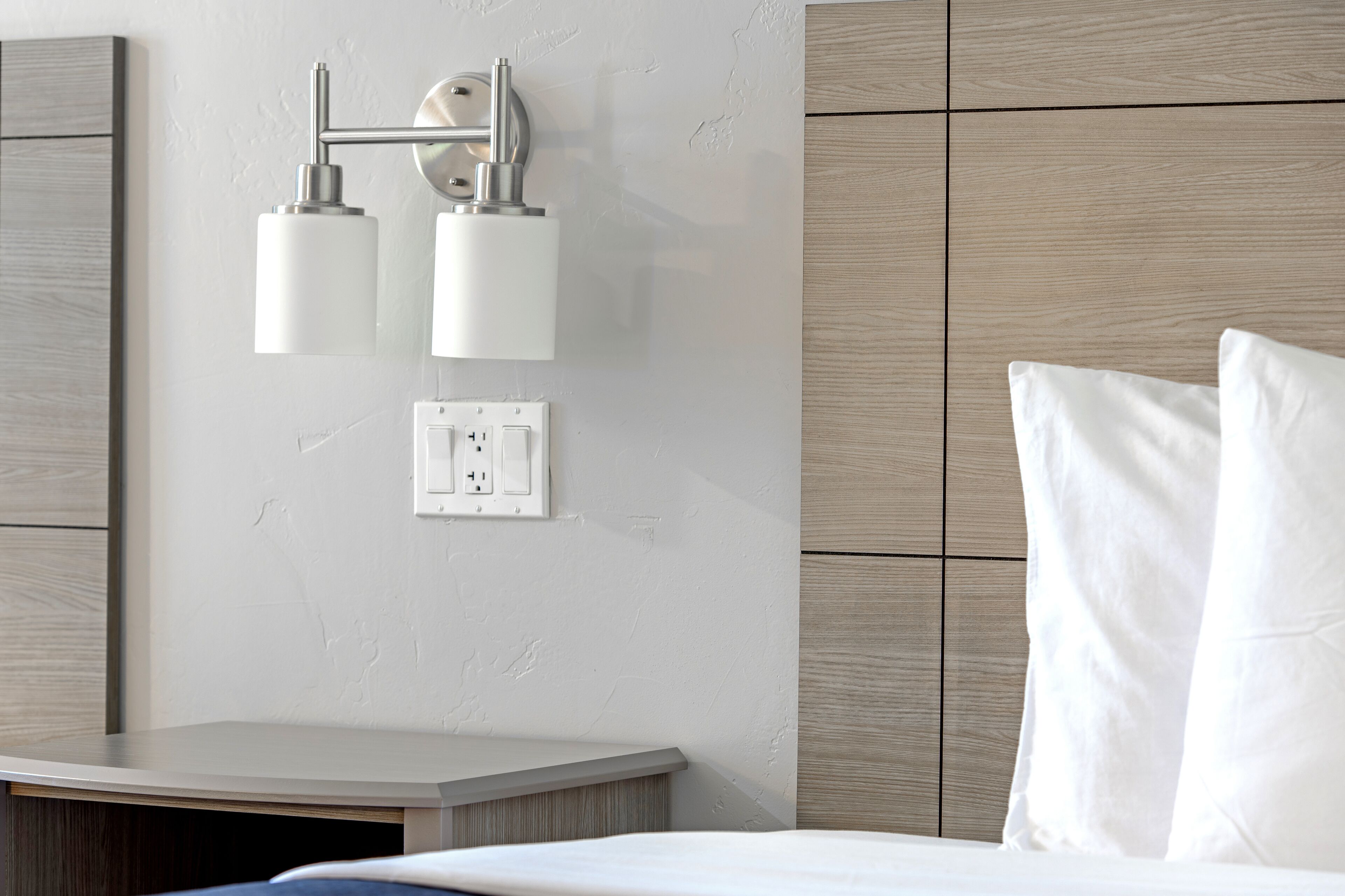 Een moderne hotelkamer met een houten hoofdeinde, een nachtkastje, een wandlamp en een stopcontact.