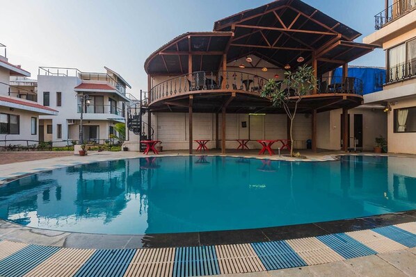 A swimming pool with a covered area and red tables, surrounded by buildings. - Sia Resort And Spa (Alibag)