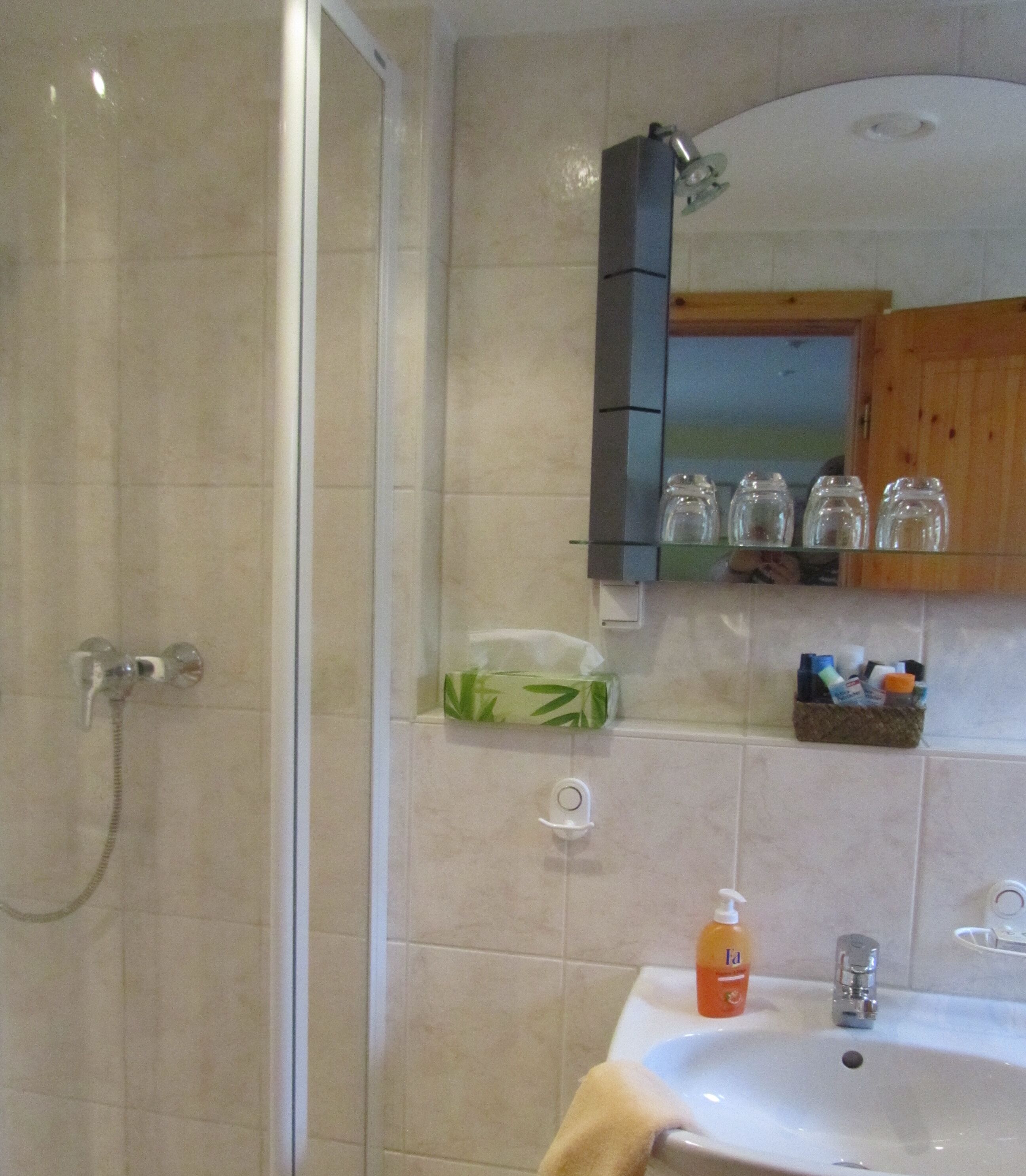 Een badkamer met een douche, een wastafel en een spiegel erboven. Er staan toiletartikelen op de wastafel en een plank met potjes boven de spiegel.